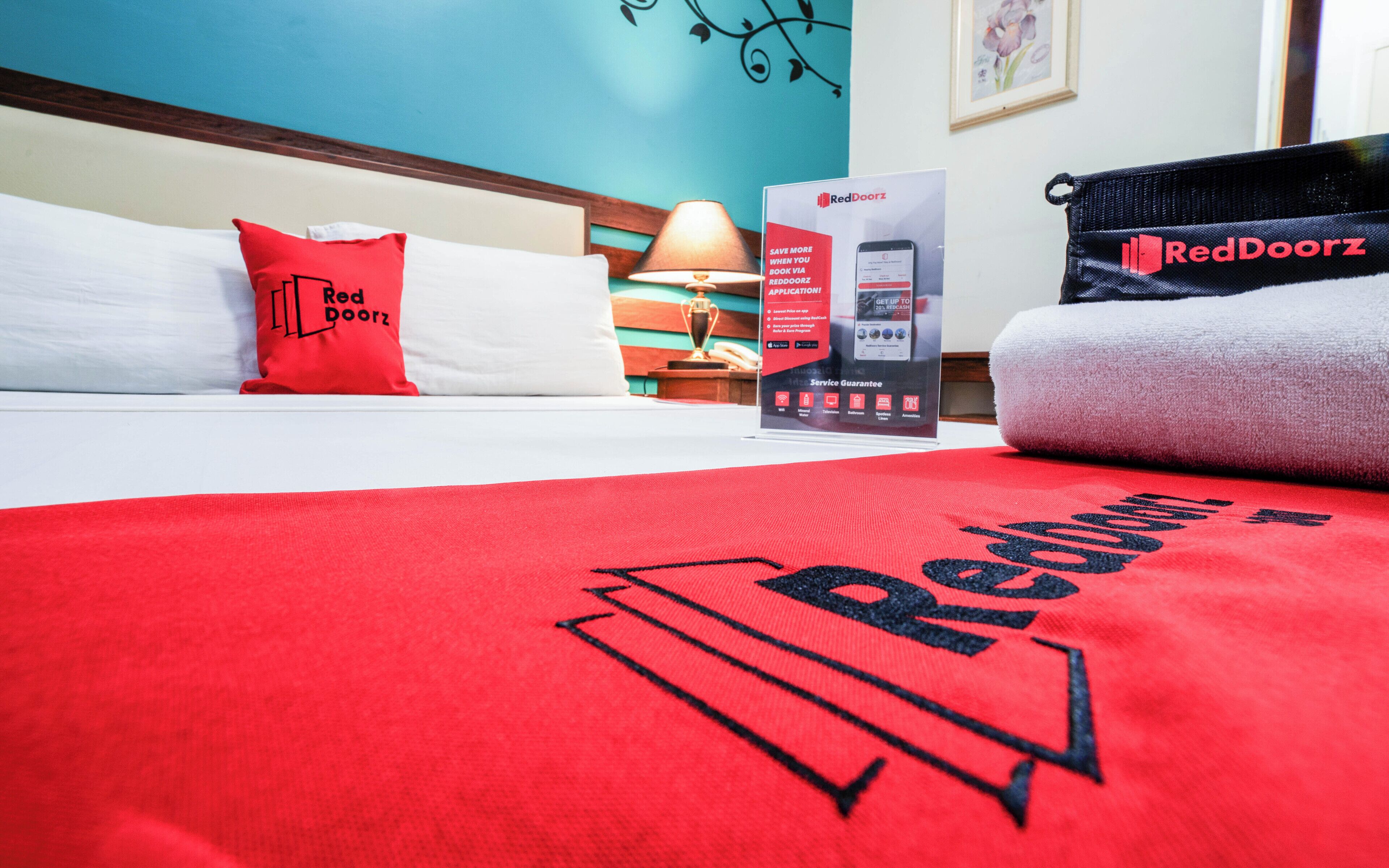 A hotel room with a bed, a RedDoorz branded pillow, a RedDoorz brochure, and a RedDoorz branded towel.