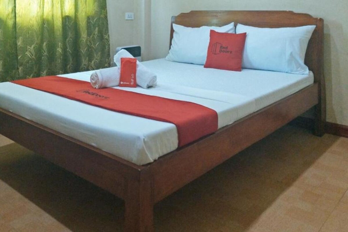 Un letto rifatto con un copriletto rosso e cuscini, una testiera in legno e una tenda verde sullo sfondo.