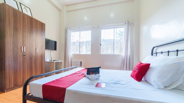 A neatly made bed with a red blanket, a pillow, and a folded towel. There is a wooden wardrobe and a television on the wall.