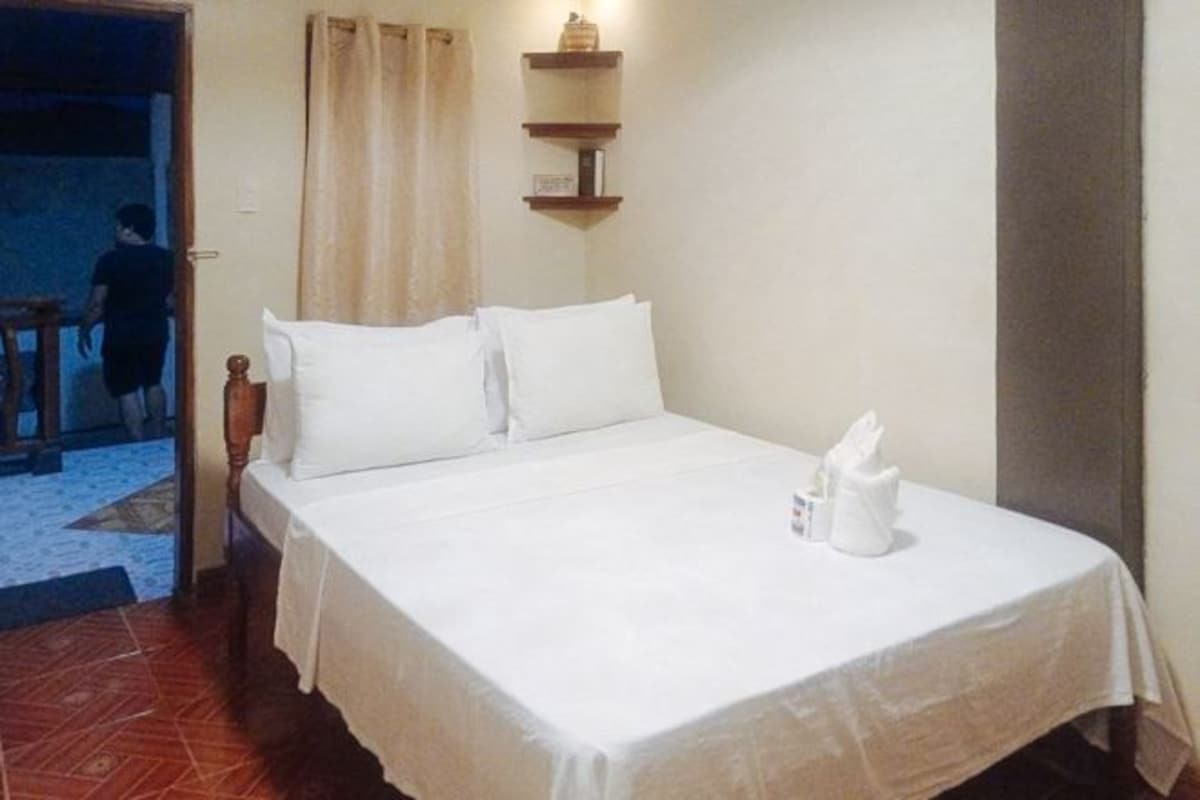 A neatly made bed with white linens and pillows, a small bedside table with a bottle and a glass, and a wall-mounted shelf with a few items.