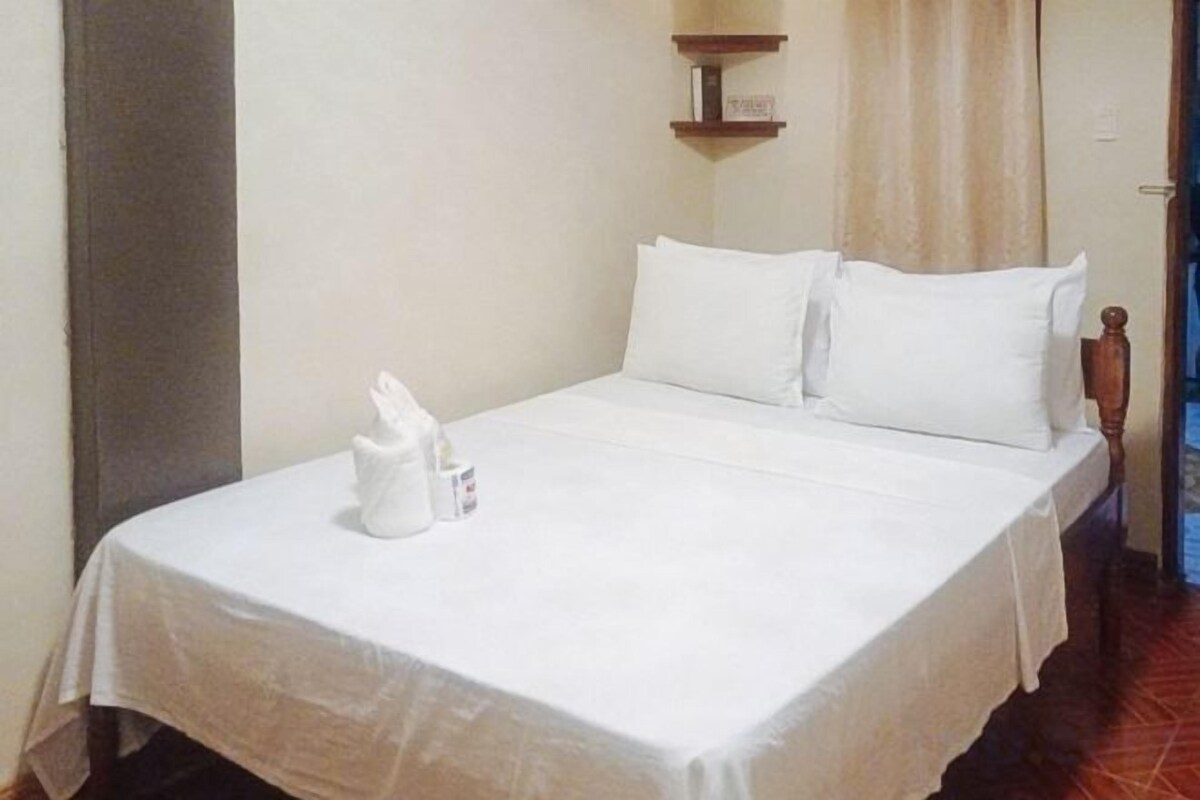 A neatly made bed with white linens and pillows, a small table with a bottle and a glass, and a wooden nightstand.