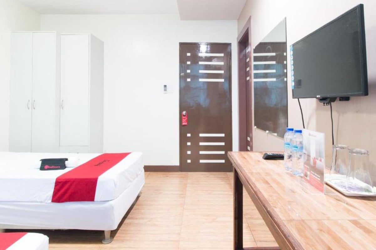 Une chambre d’hôtel avec deux lits, une table en bois sur laquelle se trouvent des bouteilles d’eau, une télévision et une porte avec un panneau rouge indiquant la sortie de secours.