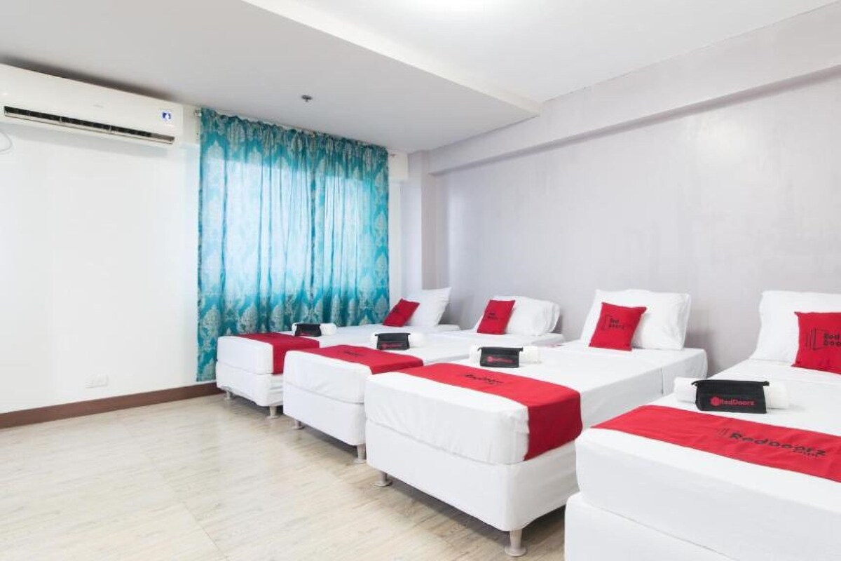 Habitación de hotel con cuatro camas individuales, cada una con cabecera y almohada roja, y manta roja. Hay un ventanal con cortinas azules, un equipo de aire acondicionado en la pared y suelo de madera.