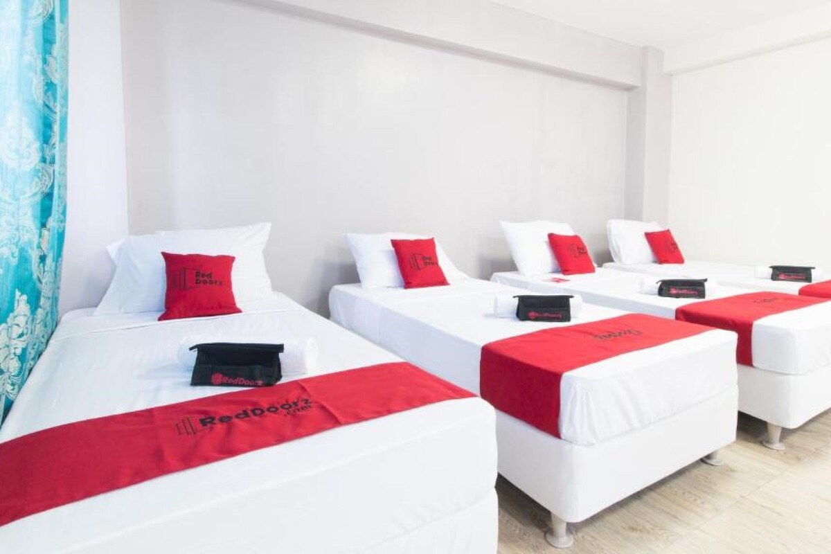 Habitación de hotel con cuatro camas, cada una con ropa de cama blanca y detalles en rojo, y una cortina azul a la izquierda.