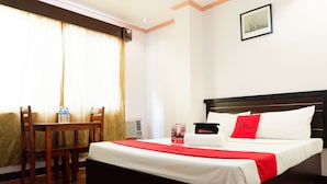 Quarto de hotel com cama, escrivaninha e cadeira. Há uma janela com cortinas, um quadro na parede e um travesseiro vermelho na cama.