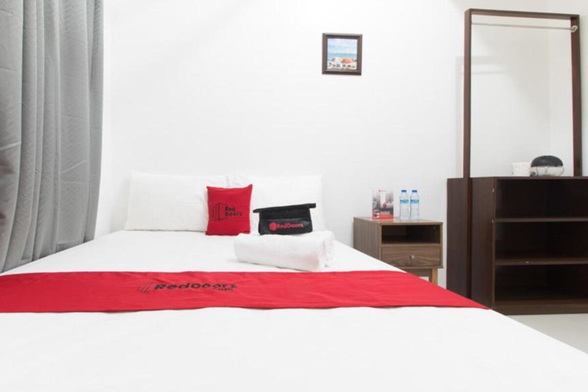 A hotel room with a neatly made bed, a red throw blanket, a pillow, a bedside table with a phone and water bottles, and a mirror.