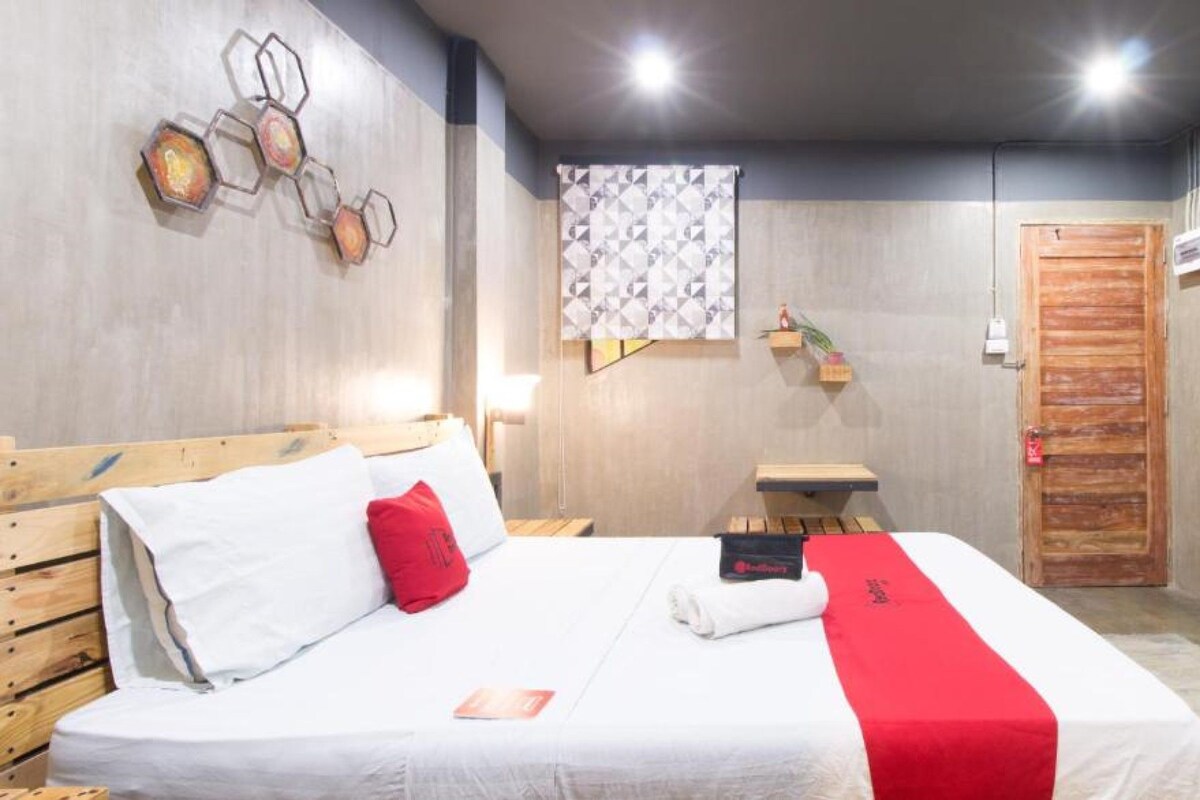 Una camera d'albergo con un letto, una testiera in legno, un cuscino rosso, una coperta bianca, una coperta rossa, un'opera d'arte appesa al muro, una tenda fantasia, un tavolino con una pianta, una porta in legno e una televisione a schermo piatto.