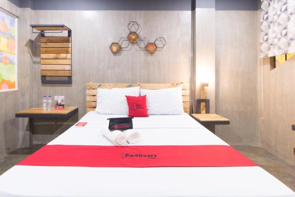 Ein Hotelzimmer mit Bett, roter Decke, RedDoorz Logo, Holzkopfstütze, Nachttisch, Wandbild und Fenster mit Blick.