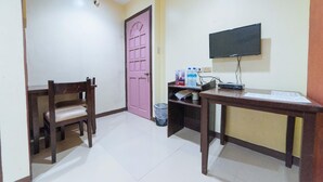 Una habitación pequeña con un escritorio de madera, una silla, una puerta rosa, un televisor montado en la pared y un cubo de basura.