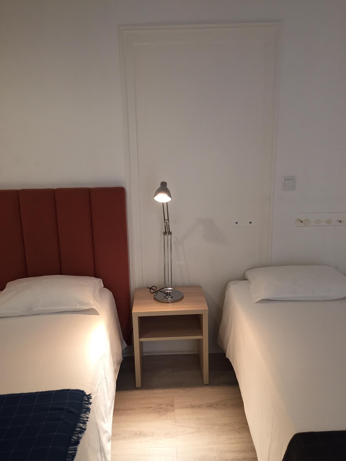 A room with two beds, a nightstand with a lamp, and a door.