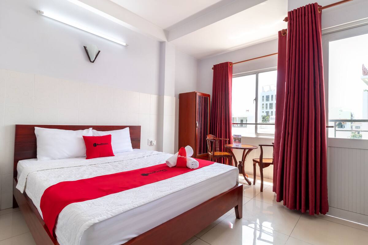 A hotel room with a large bed, a wooden headboard, a red throw blanket, a red pillow, a small table with a chair, and a window with red curtains.