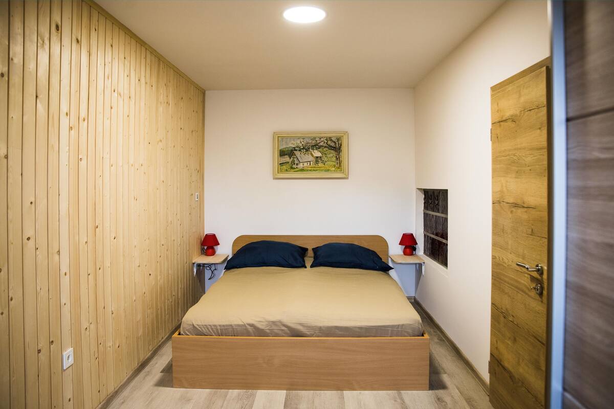 A bedroom with a wooden bed, two bedside tables with red lamps, a wooden wardrobe, and a framed picture on the wall.