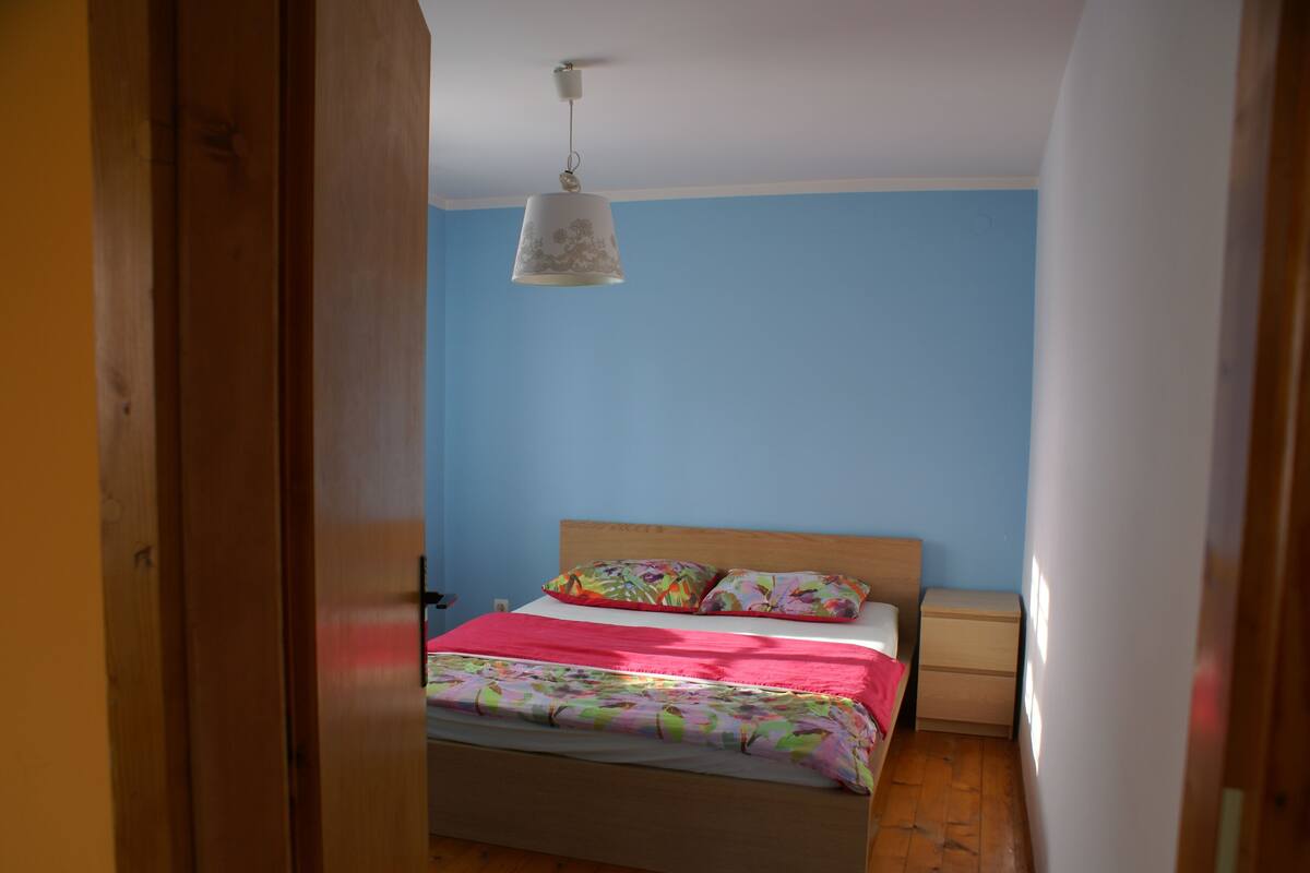 A bedroom with a bed, a nightstand, a hanging light fixture, and a wooden door.