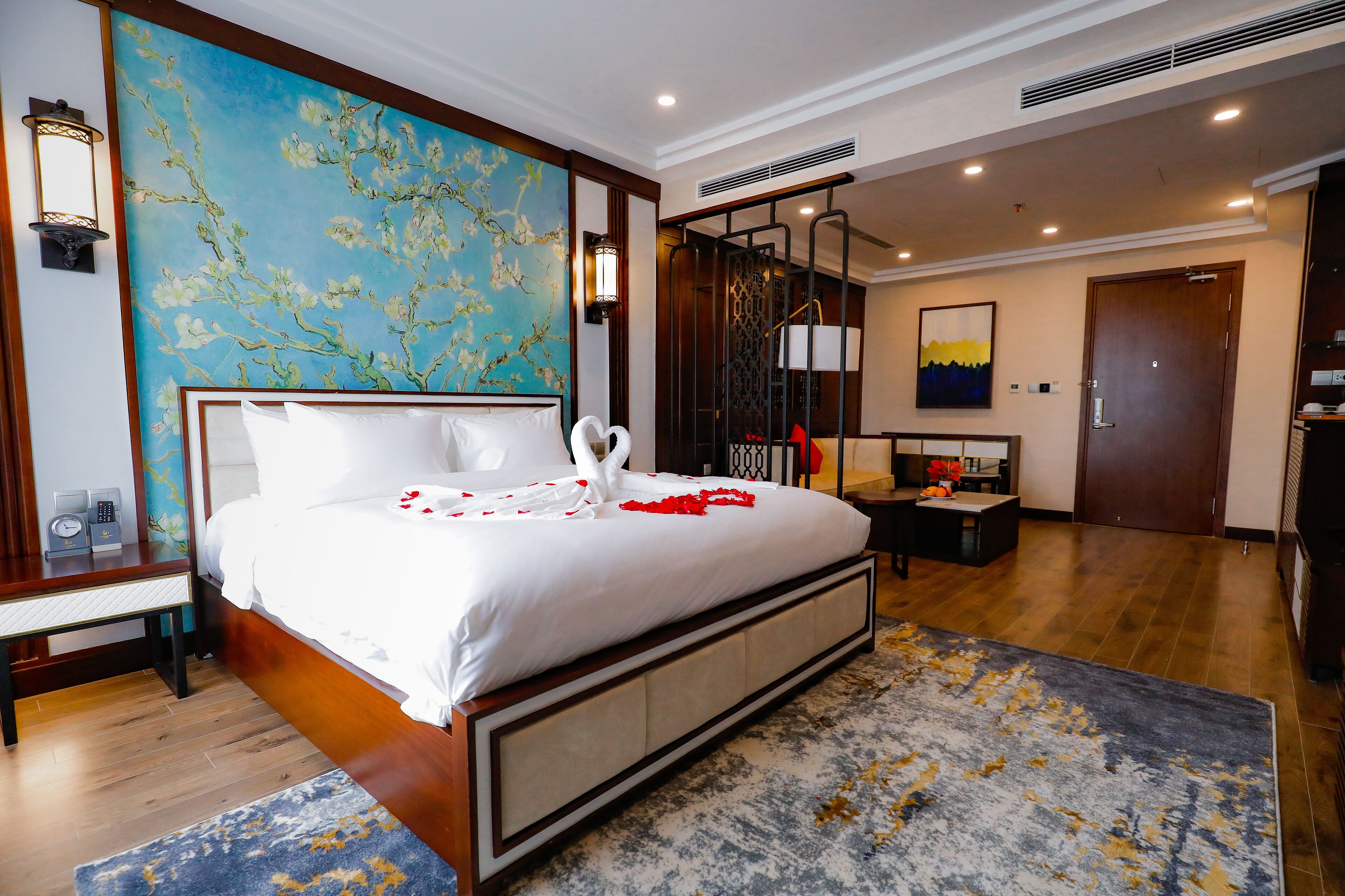 Hotelkamer met een groot bed, een houten hoofdeinde, een blauwe bloemenmuur en een modern bureau, met een deur naar een andere kamer.
