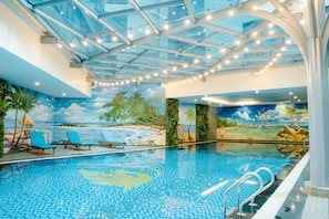 An indoor swimming pool with a glass ceiling, blue chairs, and a mural of a beach scene. - Grand Phoenix Hotel Bac Ninh (Vo Cuong)