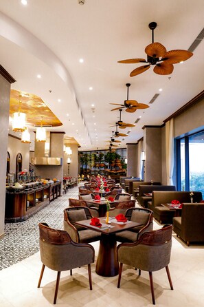 A spacious hotel dining area with multiple tables and chairs, ceiling fans, and a long counter with food and beverage options. - Grand Phoenix Hotel Bac Ninh (Vo Cuong)