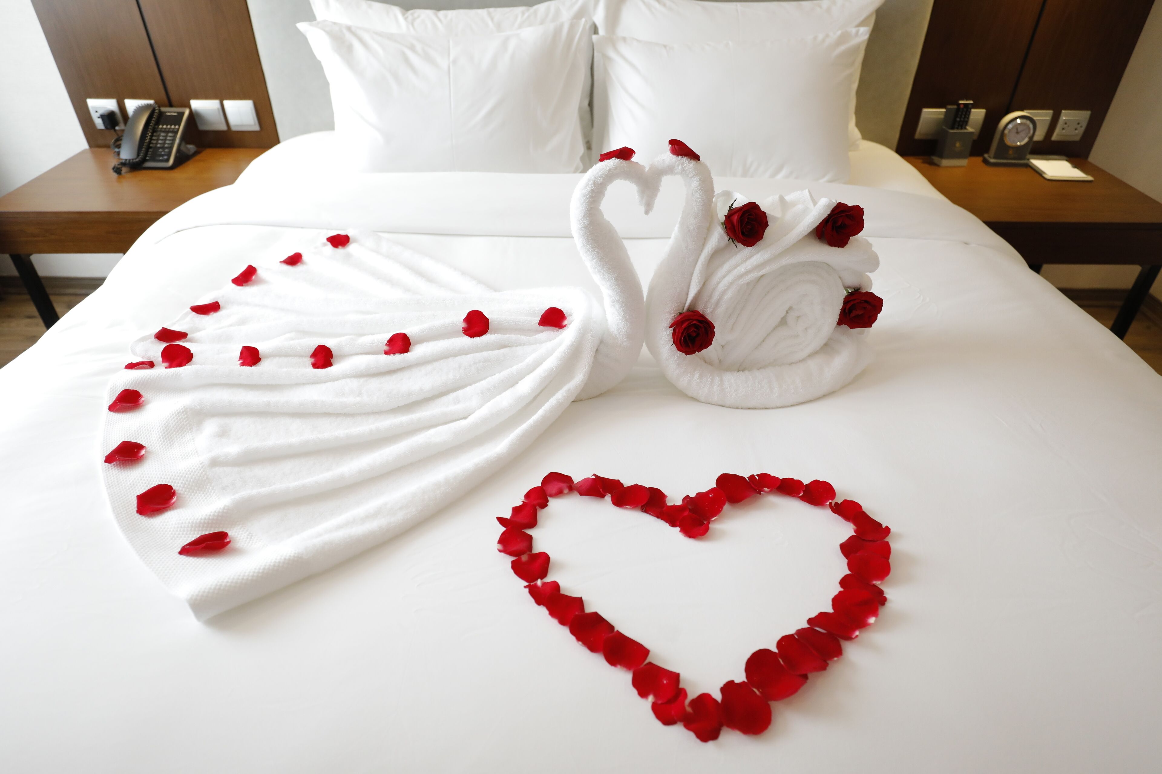 A neatly made bed with a heart-shaped arrangement of rose petals and a towel shaped like swans with roses.
