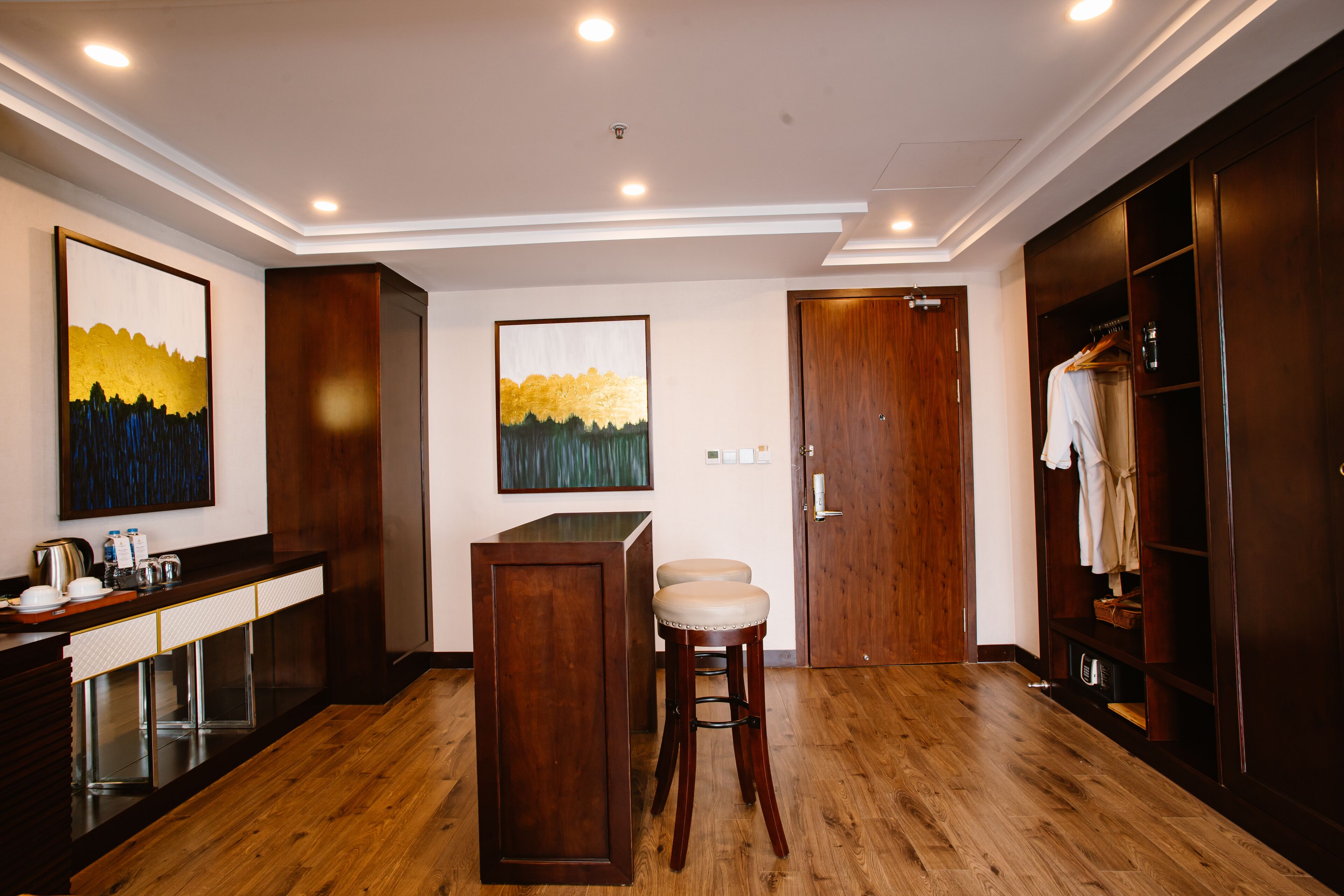 A hotel room with a wooden floor, a bar area with stools, a wardrobe, and two paintings on the wall.