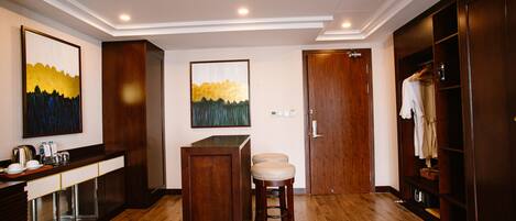 A hotel room with a wooden floor, a bar area with stools, a wardrobe, and two paintings on the wall.