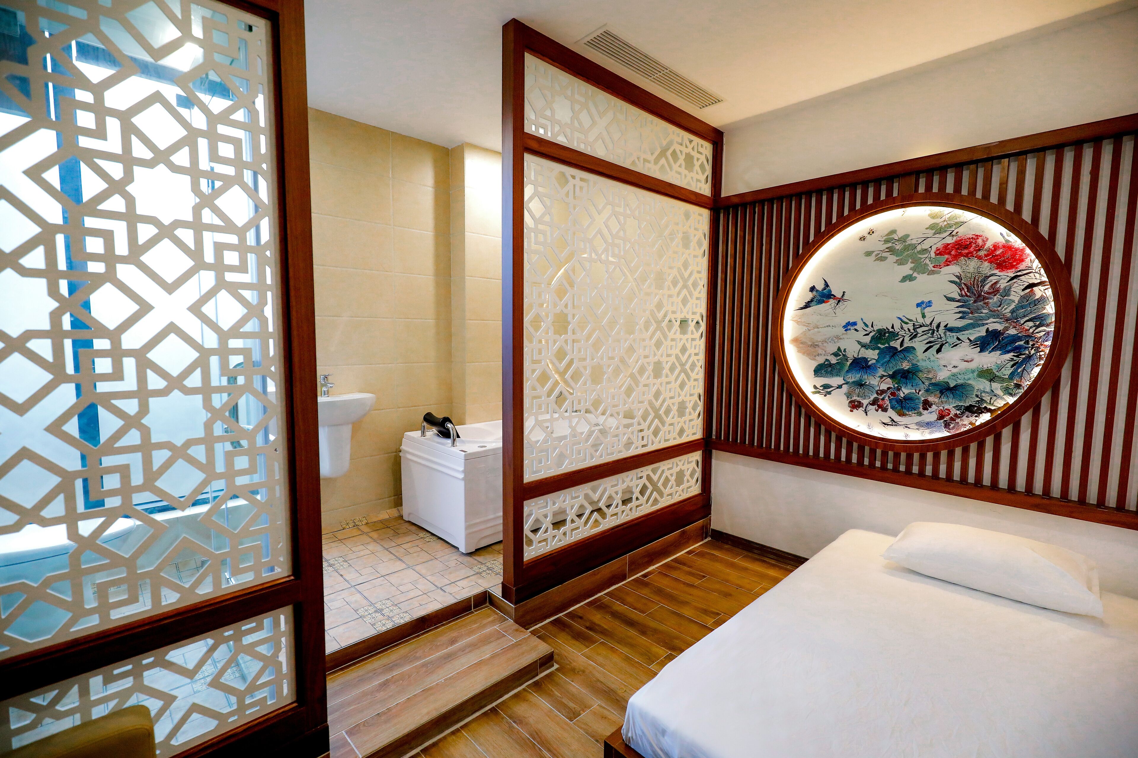 Habitación de hotel con piso de madera, cama con ropa blanca, espejo redondo con diseño floral y un inodoro con gabinete blanco.