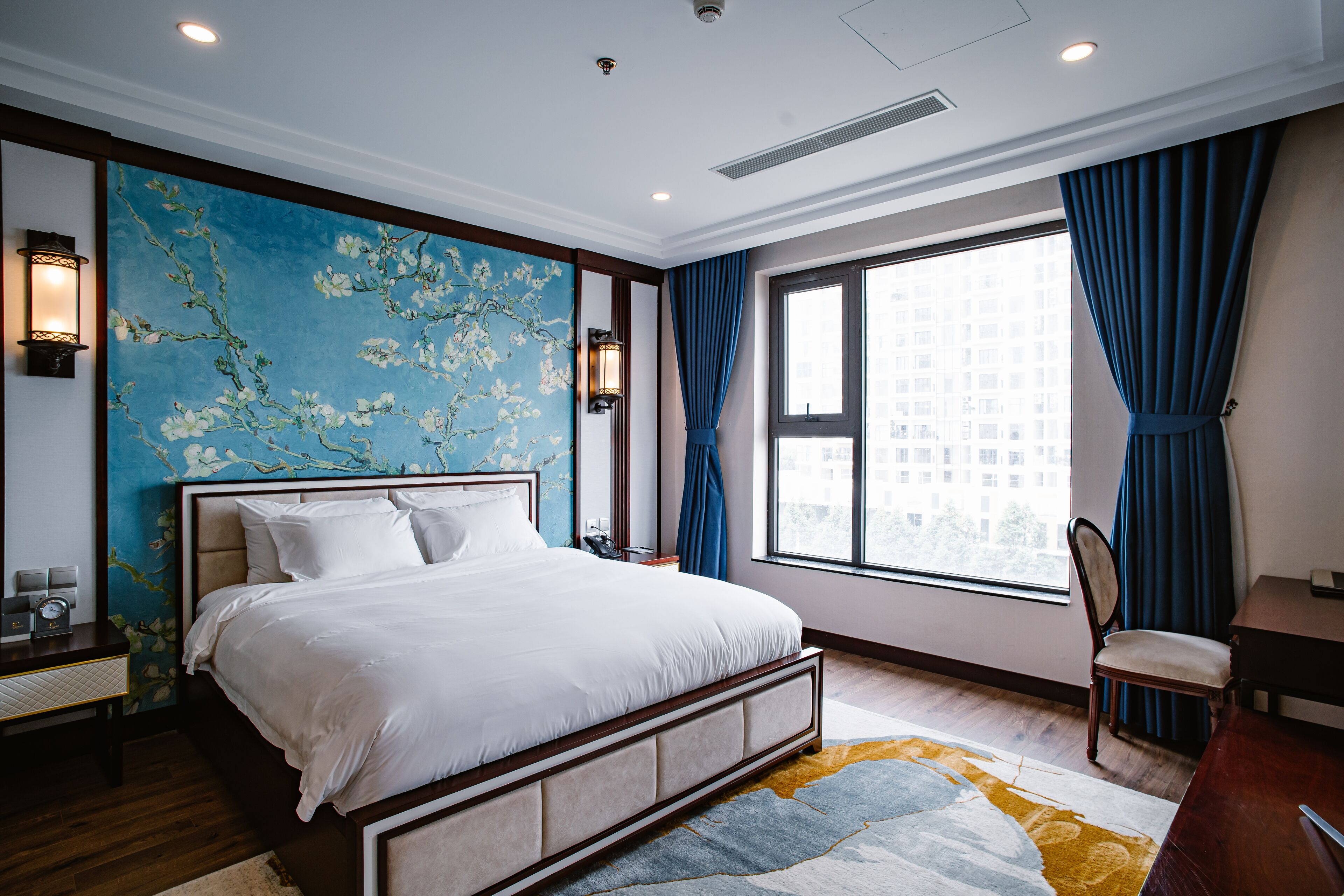 Habitación de hotel moderna con una cama grande, un escritorio y una silla. Se ve una ventana con cortinas y vistas a la ciudad.