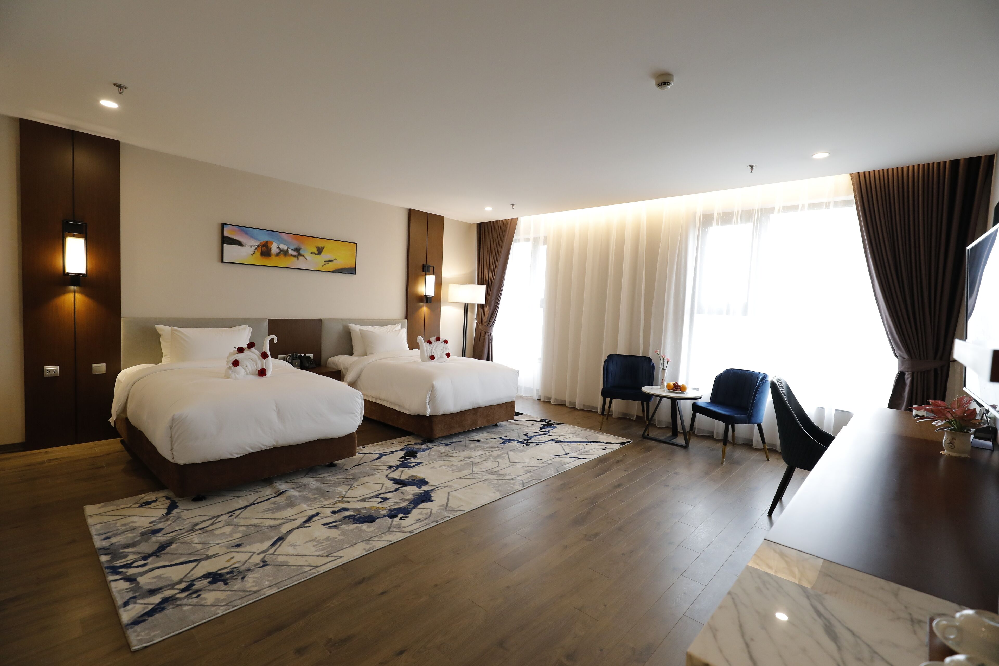 A hotel room with two beds, a dining table, a chair, a small table with a vase, and a painting on the wall.