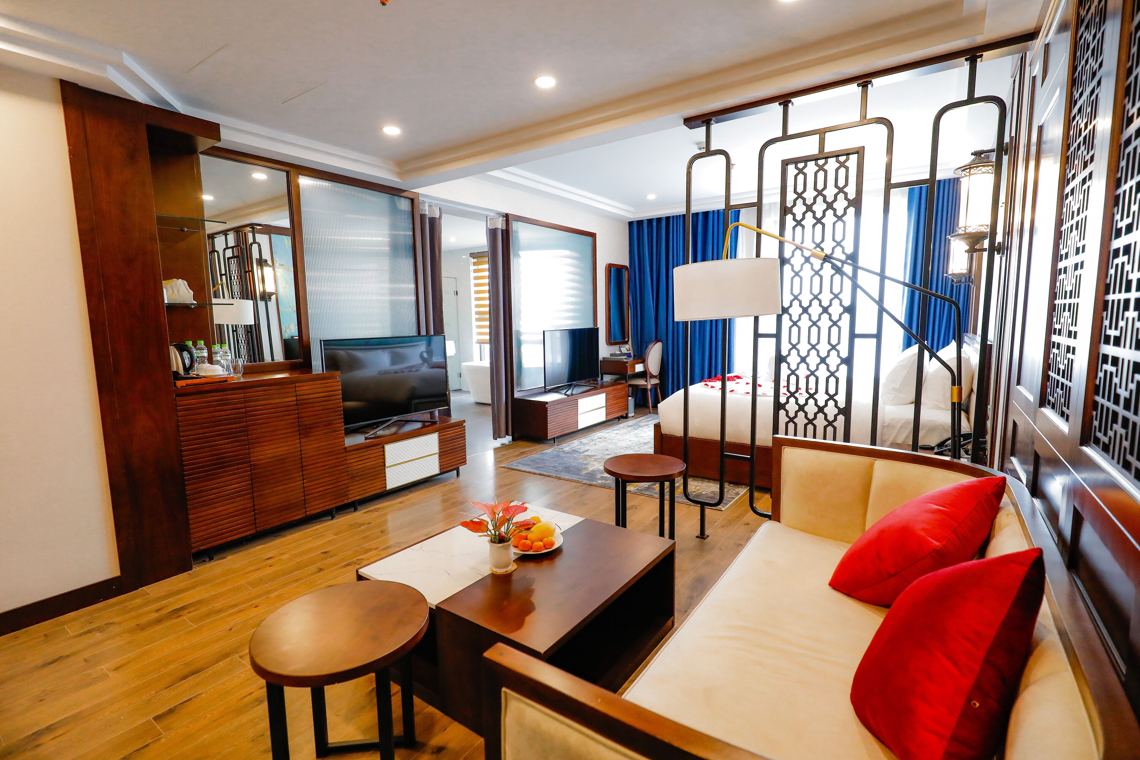 A modern hotel room with a wooden floor, a sofa with red cushions, a TV, a desk, and a large mirror.
