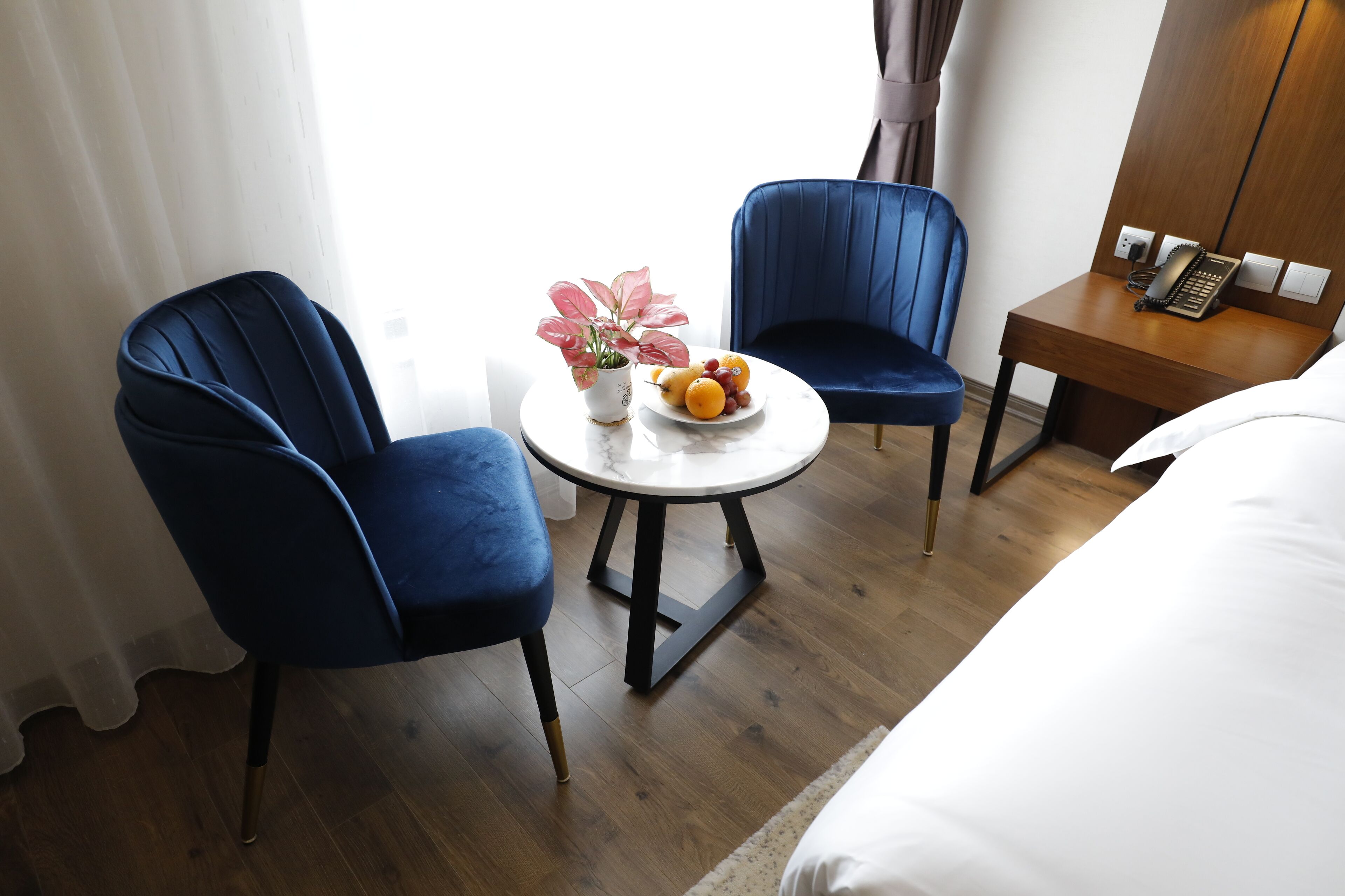 A hotel room with two blue chairs, a small round table with a fruit bowl, a potted plant, and a wooden nightstand with a phone.