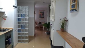 Een smalle keuken met een gasfornuis, kasten en een glazen scheidingswand. Achtergrond toont een eethoek met een tafel en stoelen.