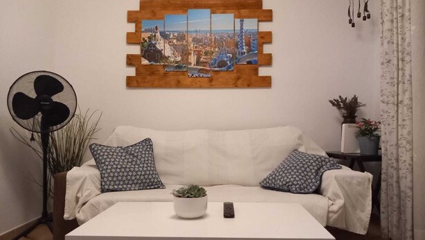 Een woonkamer met een witte bank, decoratieve kussens, een witte salontafel met een plant en afstandsbediening, een aan de muur hangend stadsgezicht, een staande ventilator en een bijzettafeltje met een potplant.