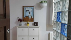 Een kamer met een witte kast, een schilderij van een vrouw bij een raam, een klok en een hangplant.