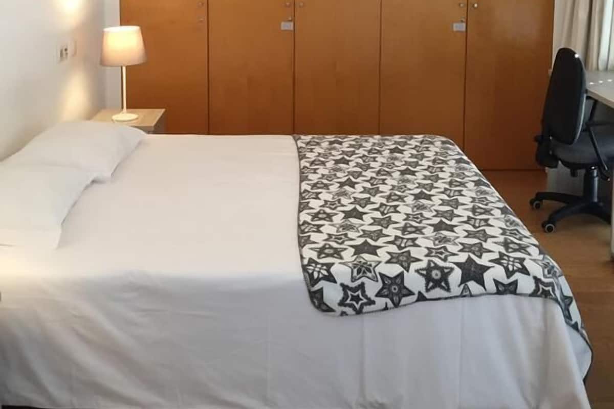 Una habitación de hotel con una cama, un escritorio, una silla, un armario y una lámpara colgante.