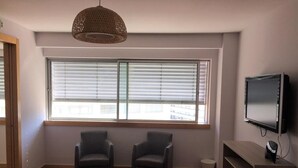 A modern living room with a large window, two armchairs, a wooden floor, a TV, and a hanging light fixture.