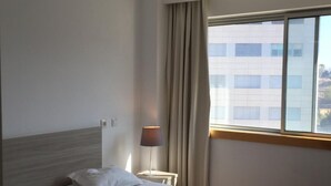 A hotel room with two beds, a lamp, a window with curtains, and a view of a building outside.