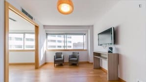 A spacious room with wooden flooring, two armchairs, a television, and a large window with a view of buildings.