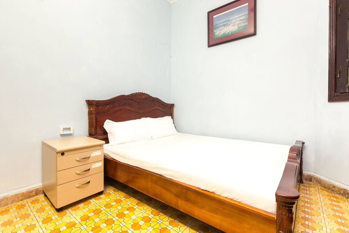 A bedroom with a wooden bed, a small bedside table with drawers, and a tiled floor.