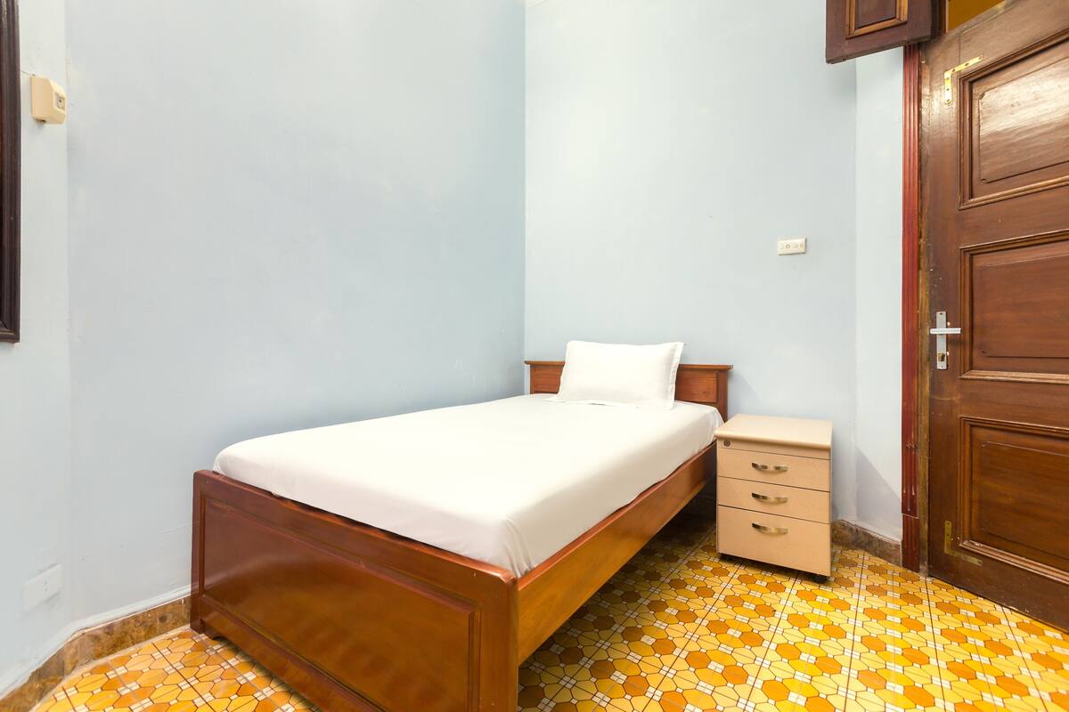 A single bed with a wooden frame and a white mattress, a small bedside table with drawers, and a door with a wooden frame.