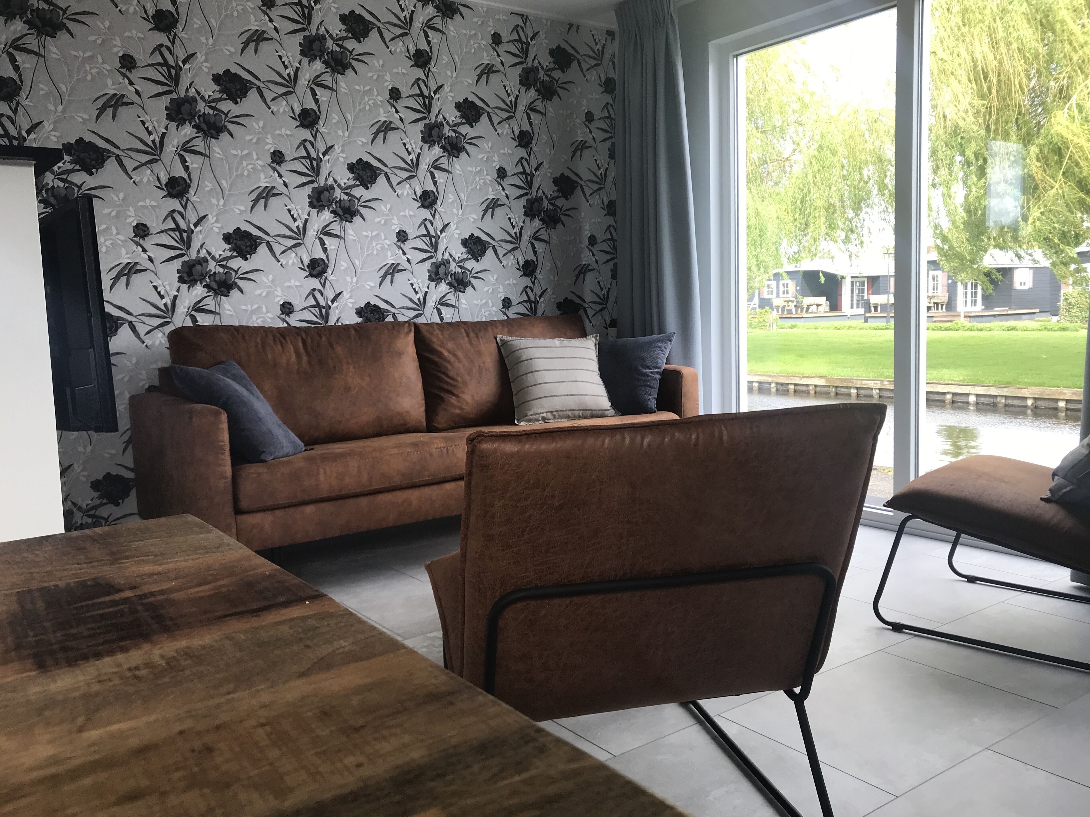 Una sala de estar moderna con un sofá marrón, una mesa de comedor de madera y vistas a un paisaje verde a través de amplios ventanales.
