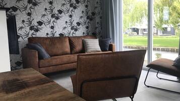 Una sala de estar moderna con un sofá marrón, una mesa de comedor de madera y vistas a un paisaje verde a través de amplios ventanales.