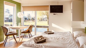 A modern hotel room with a large bed, a desk, and chairs. There is a view of a garden and a pool outside the window.