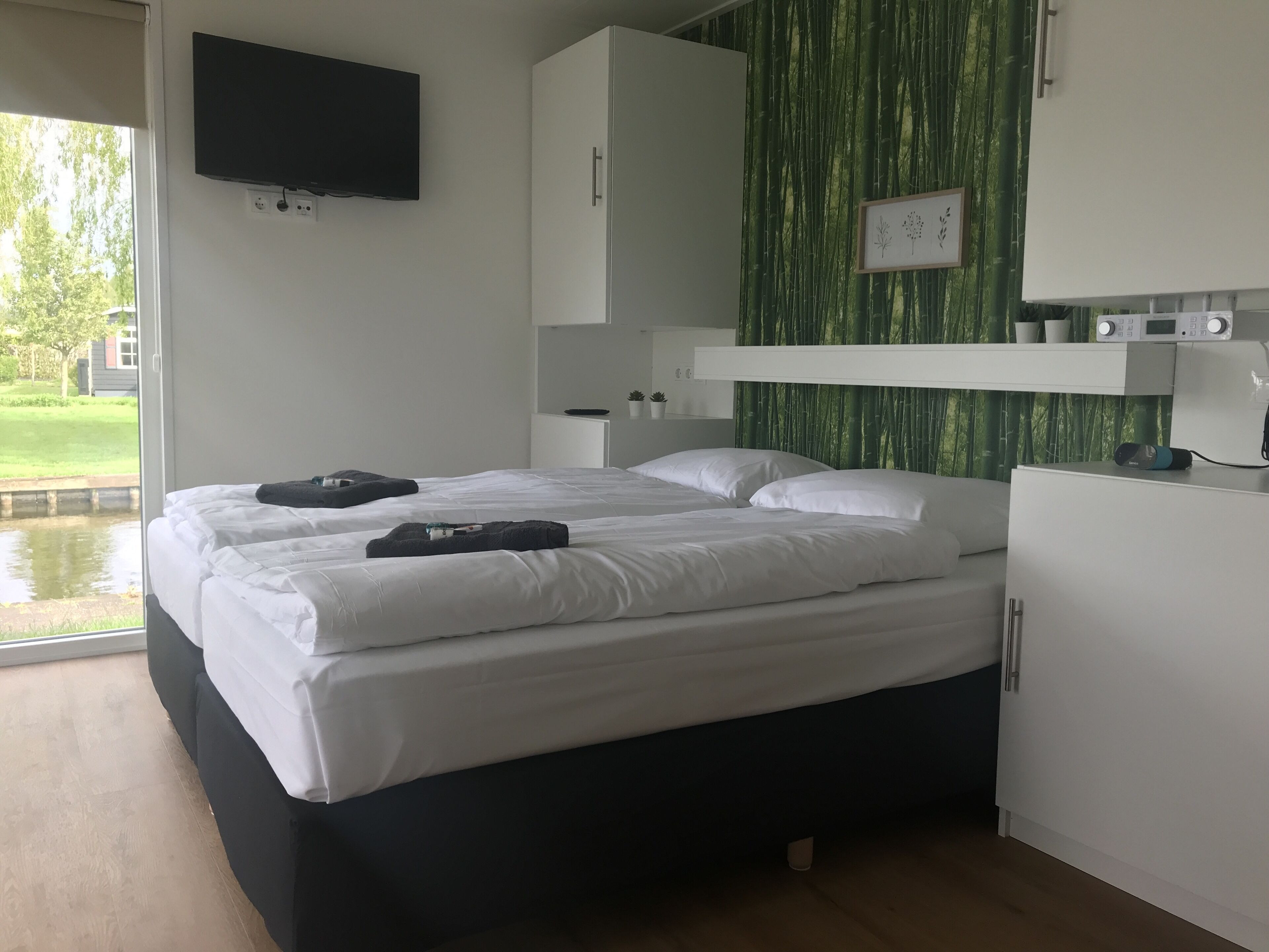 A modern bedroom with a bed, a TV mounted on the wall, a small cabinet, and a window with a view of greenery outside.