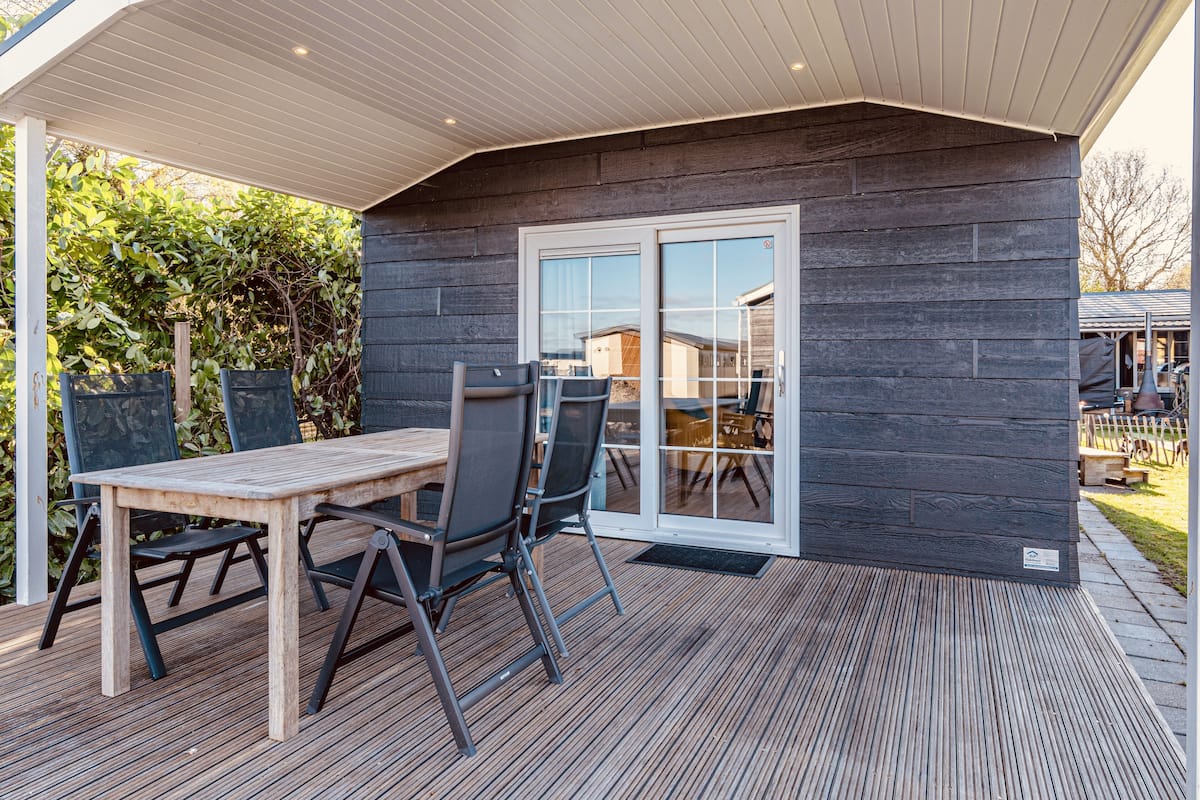 Una terraza de madera con una mesa y sillas, un porche cubierto y un ventanal corredizo que da a un espacio exterior techado.