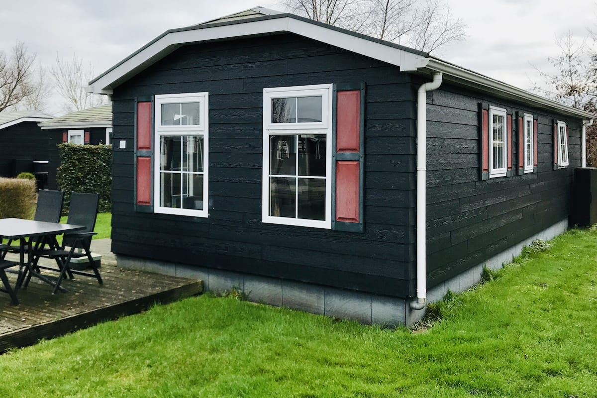 Una casa pequeña de color oscuro con ventanas de marcos blancos y contraventanas rojas, un patio con mesa y sillas, y un césped bien cuidado.