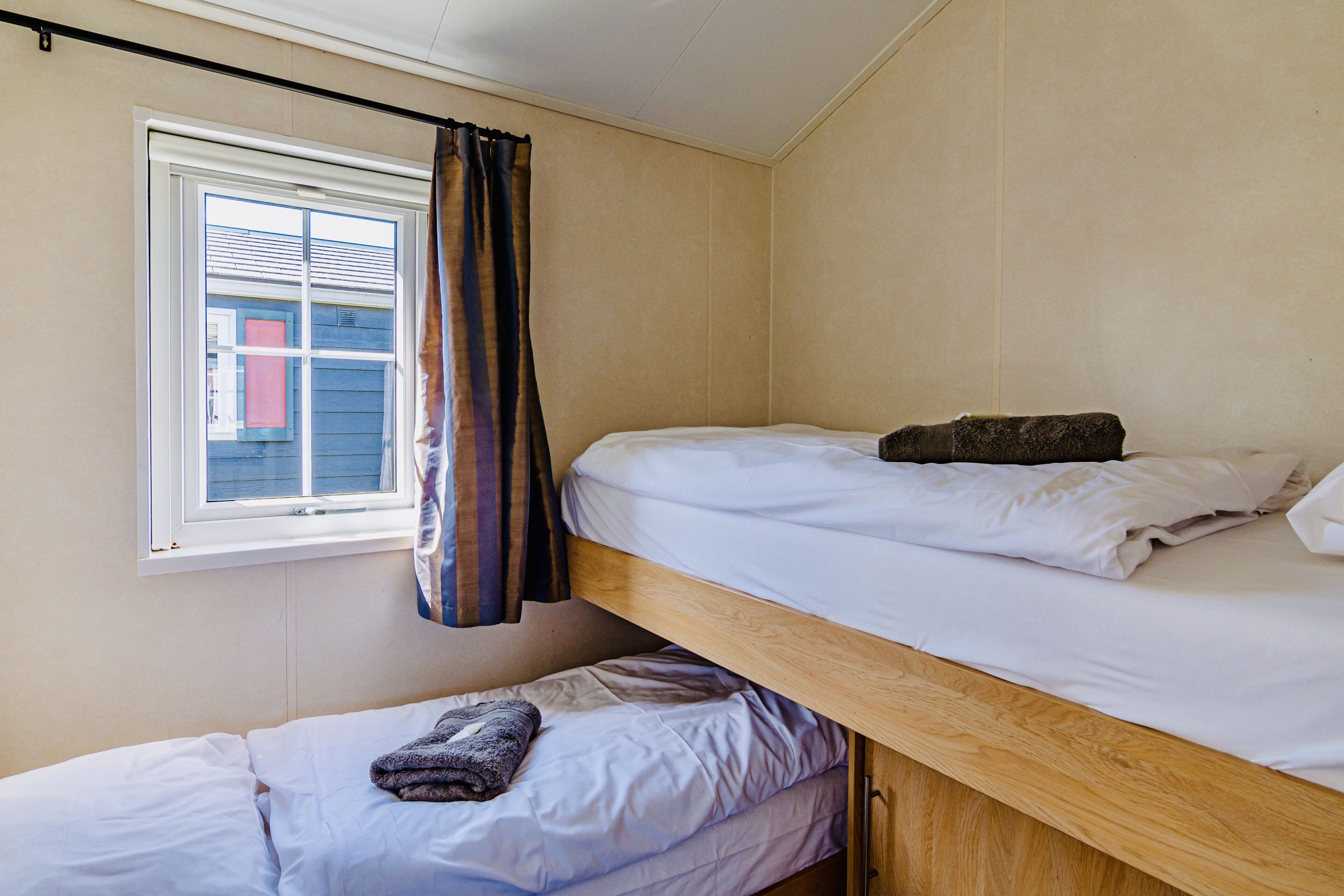 Quarto estilo dormitório com beliches, janela com cortinas e vista para um edifício.