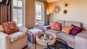 A living room with a sofa, armchair, and a coffee table. There are curtains, a floor lamp, and wall decor.