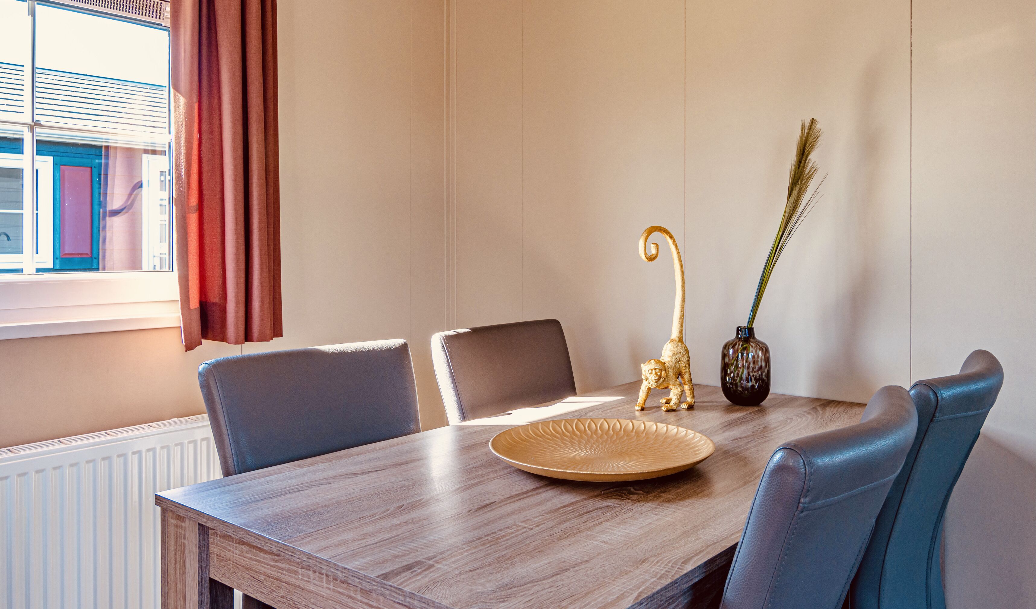 Un coin repas comprenant une table en bois, quatre chaises, une assiette décorative, un vase avec des plantes et une fenêtre avec des rideaux.