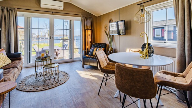 A spacious living room with a large sliding glass door leading to a balcony, a round dining table with chairs, a sofa, and a television mounted on the wall.