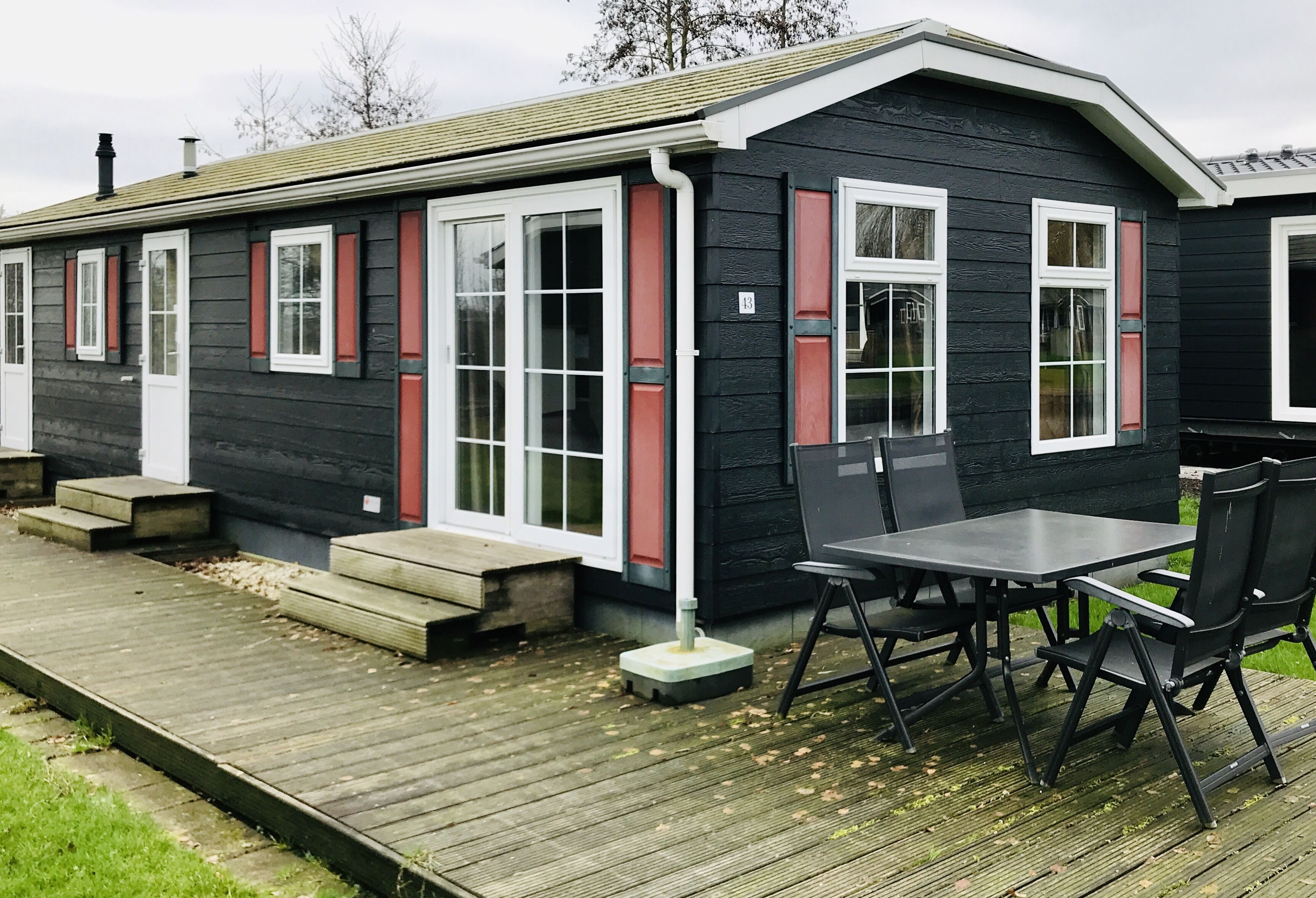A small, single-story building with a dark exterior, white-framed windows, and a wooden deck with a table and chairs.