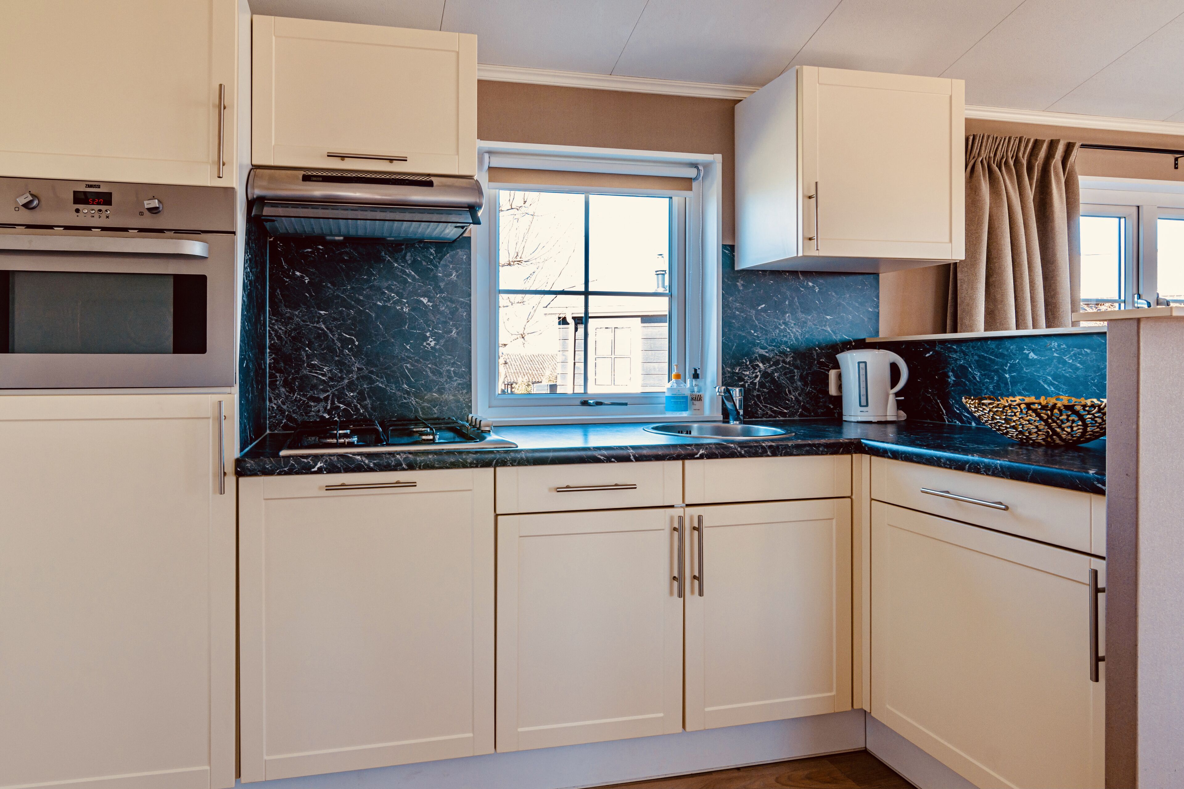 A compact kitchen with white cabinets, a built-in oven, a stovetop, and a window with curtains.