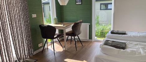 A room with a bed, a desk, and chairs. There is a window with a view of greenery outside.